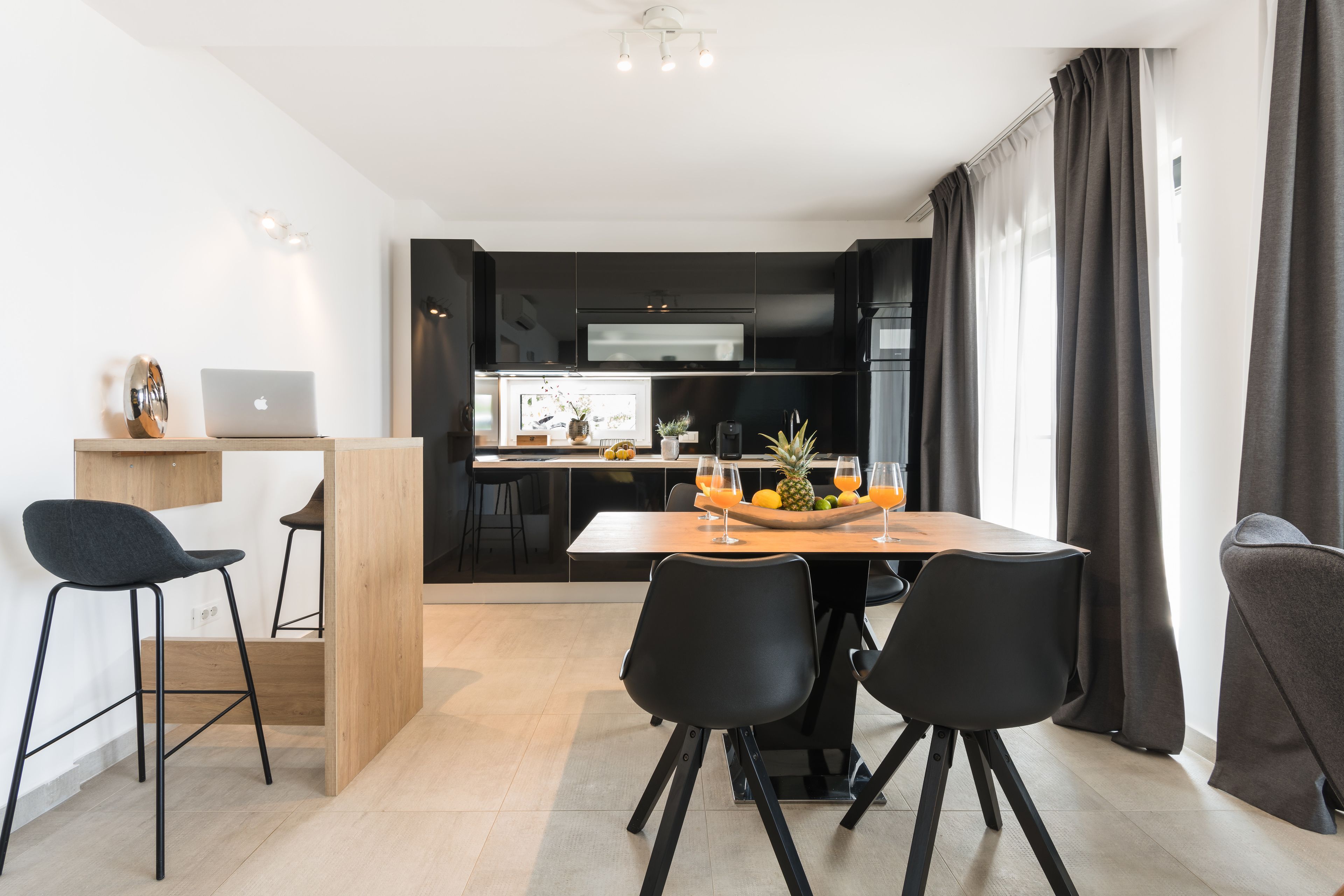Una cocina moderna con armarios oscuros, una mesa de comedor de madera con sillas negras, y una computadora portátil sobre una mesita auxiliar.
