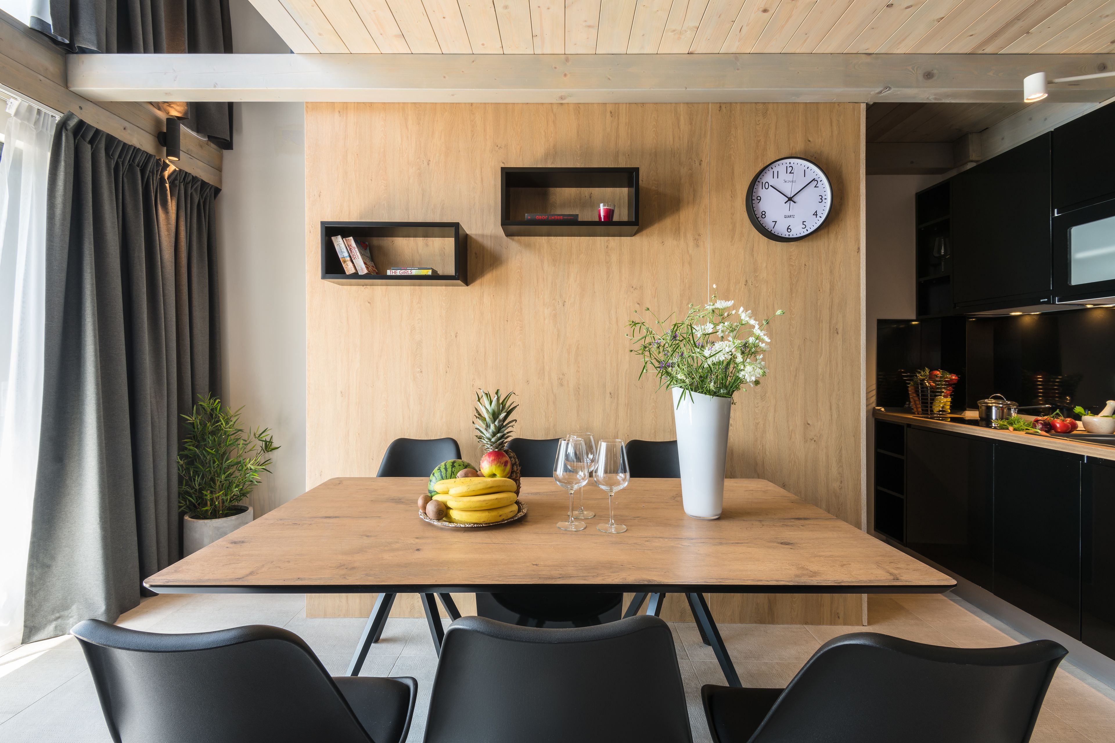 A modern dining area with a wooden table, black chairs, a clock, and a potted plant.