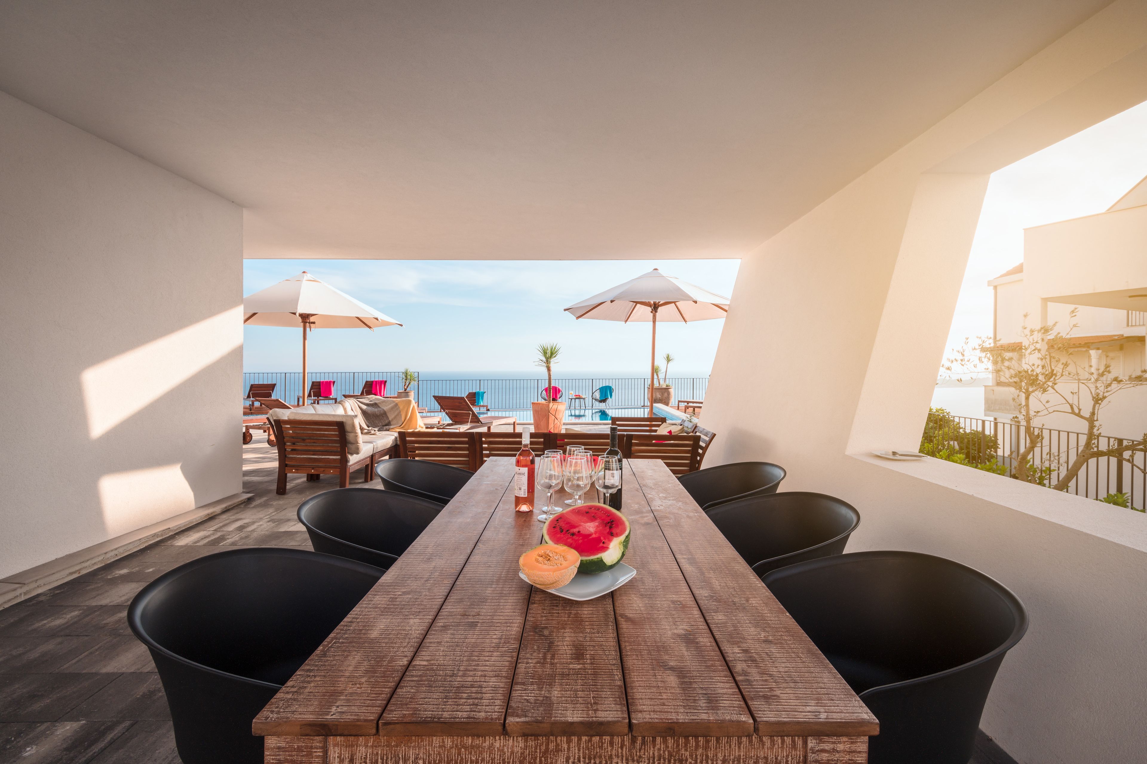 A wooden table with black chairs and a watermelon centerpiece, overlooking a beach.