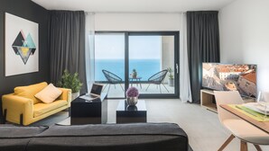 Una habitación de hotel moderna con sofá, mesa de centro, laptop, televisor y balcón con vista al mar.
