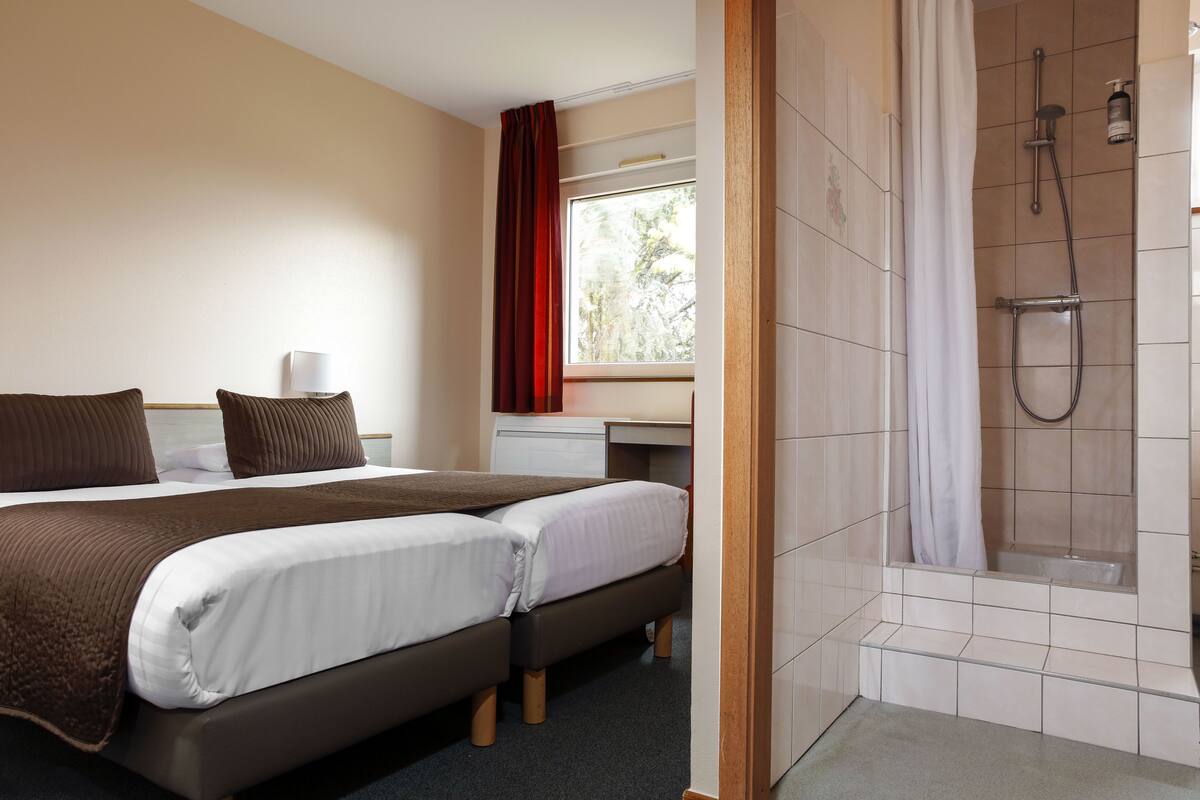 A hotel room with a bed, a desk, a shower, and a window with curtains.