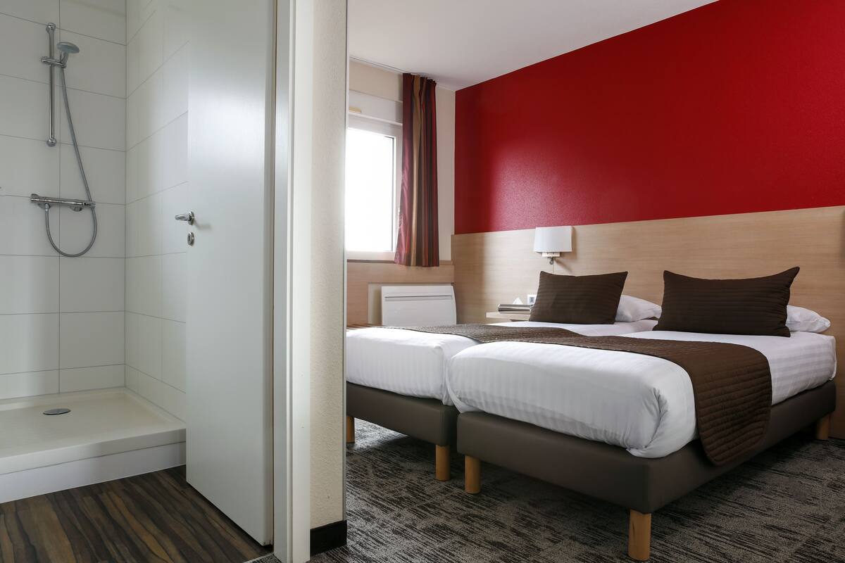A hotel room with a shower, two beds, a red accent wall, and a window with curtains.