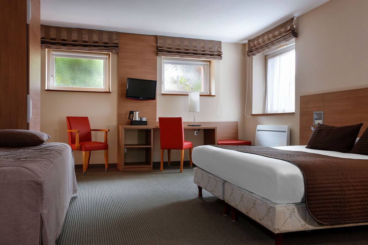 A hotel room with a bed, two red chairs, a desk with a television, and a window with curtains.