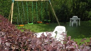 A garden with a swing set, a white plastic chair, and purple-leafed plants. - Hôtel La Clef Des Champs (Biesheim)