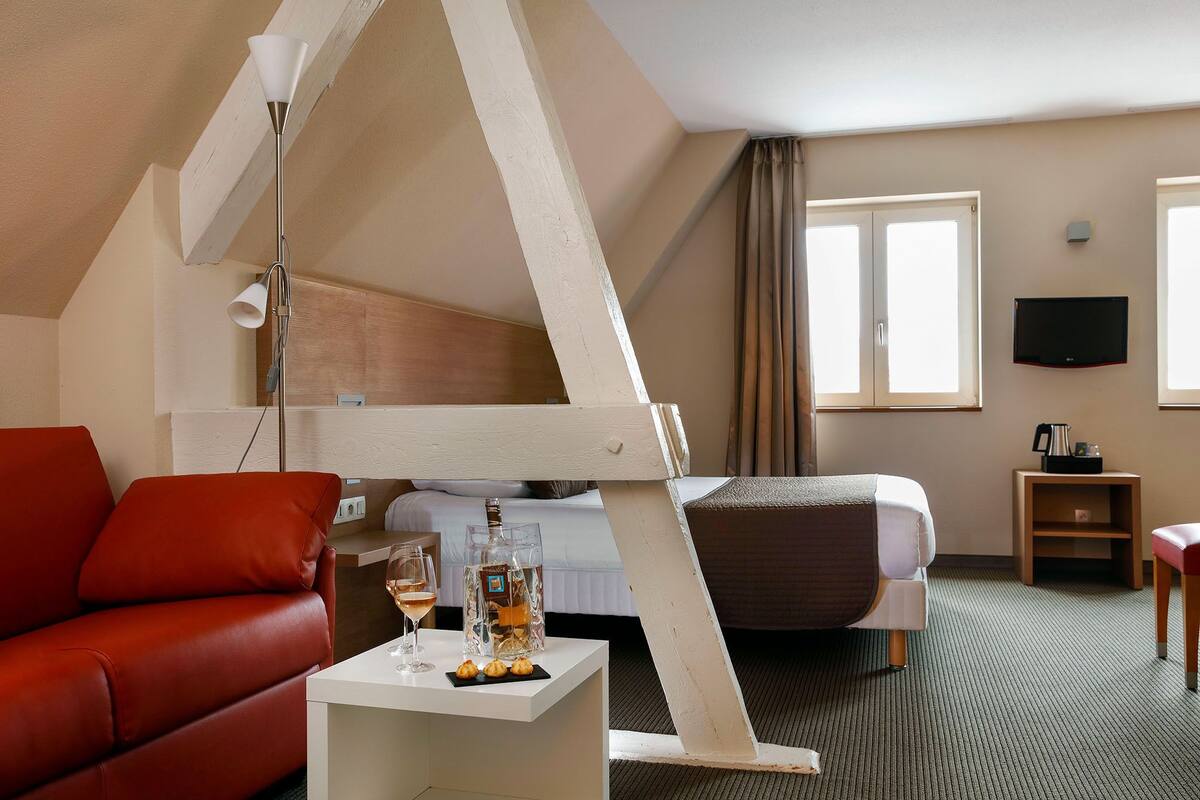 A hotel room with a bed, a sofa, a small table with a bottle and glasses, a TV, and a window.