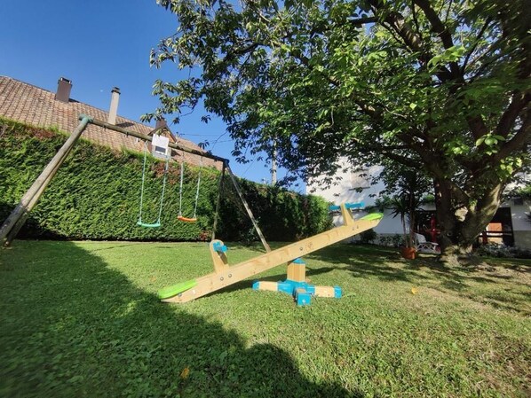 A wooden seesaw in a yard with a tree and a hedge in the background.