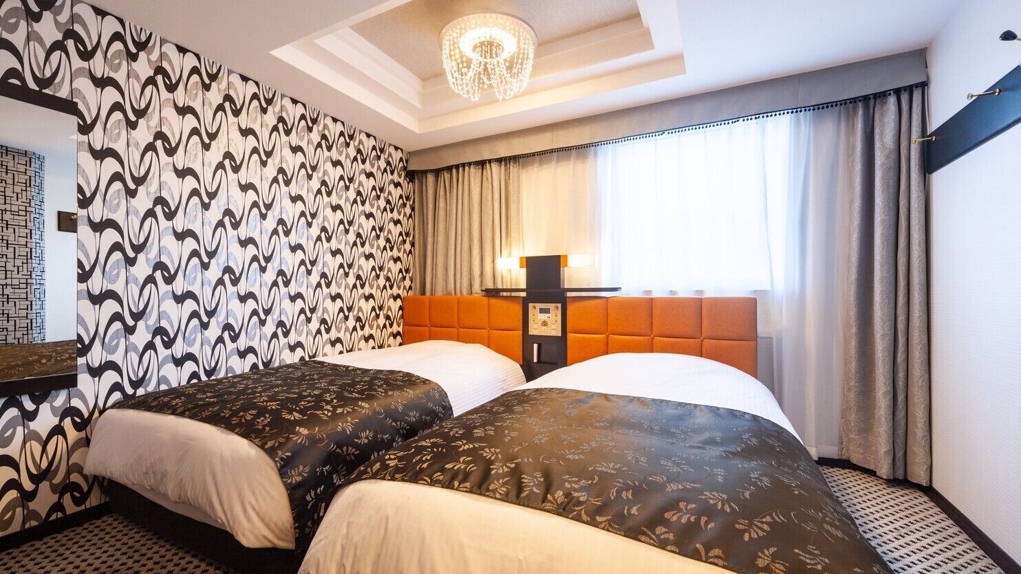 Een hotelkamer met twee bedden, een behang met patroon, een kroonluchter, een nachtkastje en een raam met gordijnen.
