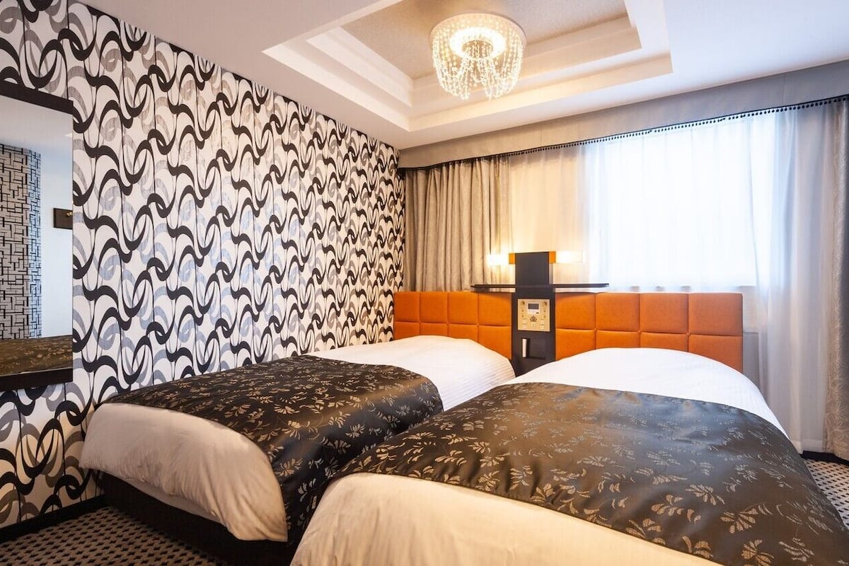 A hotel room with two beds, a patterned wall, a chandelier, a nightstand, and a window with curtains.