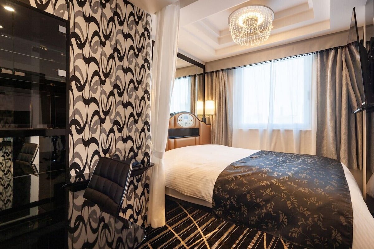 A hotel room with a bed, a desk, a chair, a chandelier, and curtains.