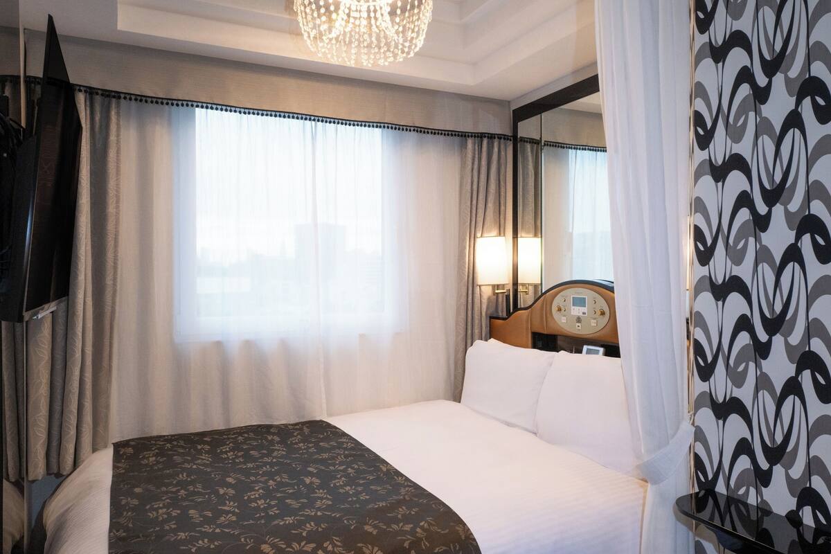 A hotel room with a large bed, a television, a chandelier, and a window with curtains.