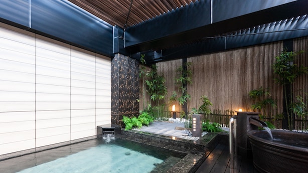 Un área de piscina exterior moderna con muelle de madera, una cascada pequeña y plantas de bambú.