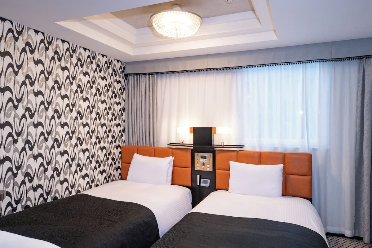 A hotel room with two beds, a control panel, a ceiling light, and patterned curtains.