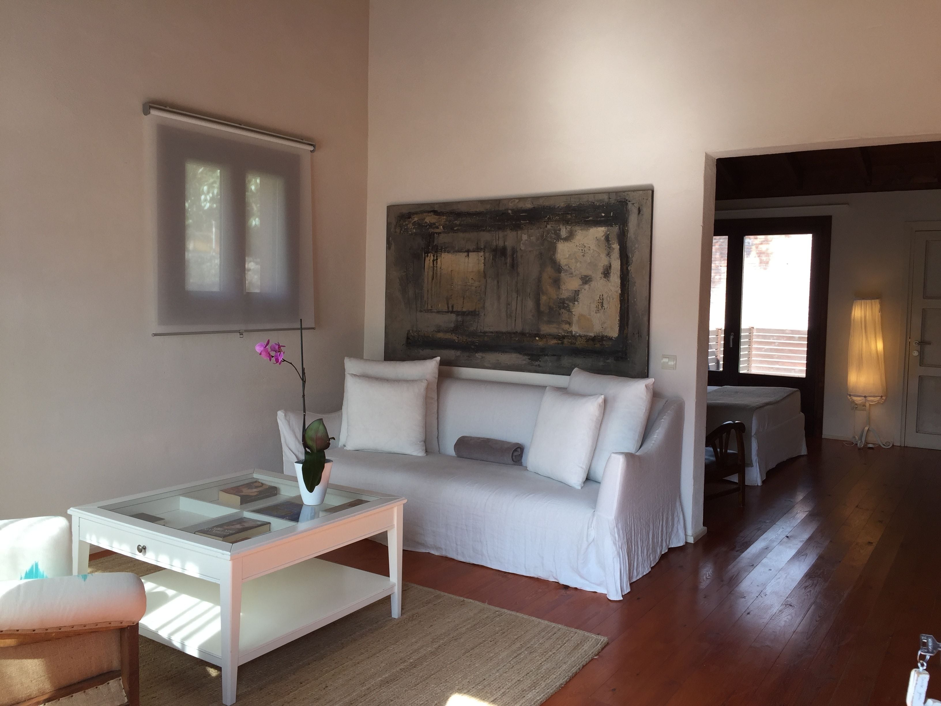 Un salón con sofá blanco, una mesa de centro de vidrio con un jarrón de flores, un cuadro en la pared y vistas al exterior a través de una puerta.
