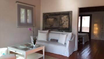 A living room with a white sofa, a glass coffee table with a vase of flowers, a painting on the wall, and a view of the outside through a door.