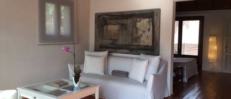 Un salón con sofá blanco, una mesa de centro de vidrio con un jarrón de flores, un cuadro en la pared y vistas al exterior a través de una puerta.