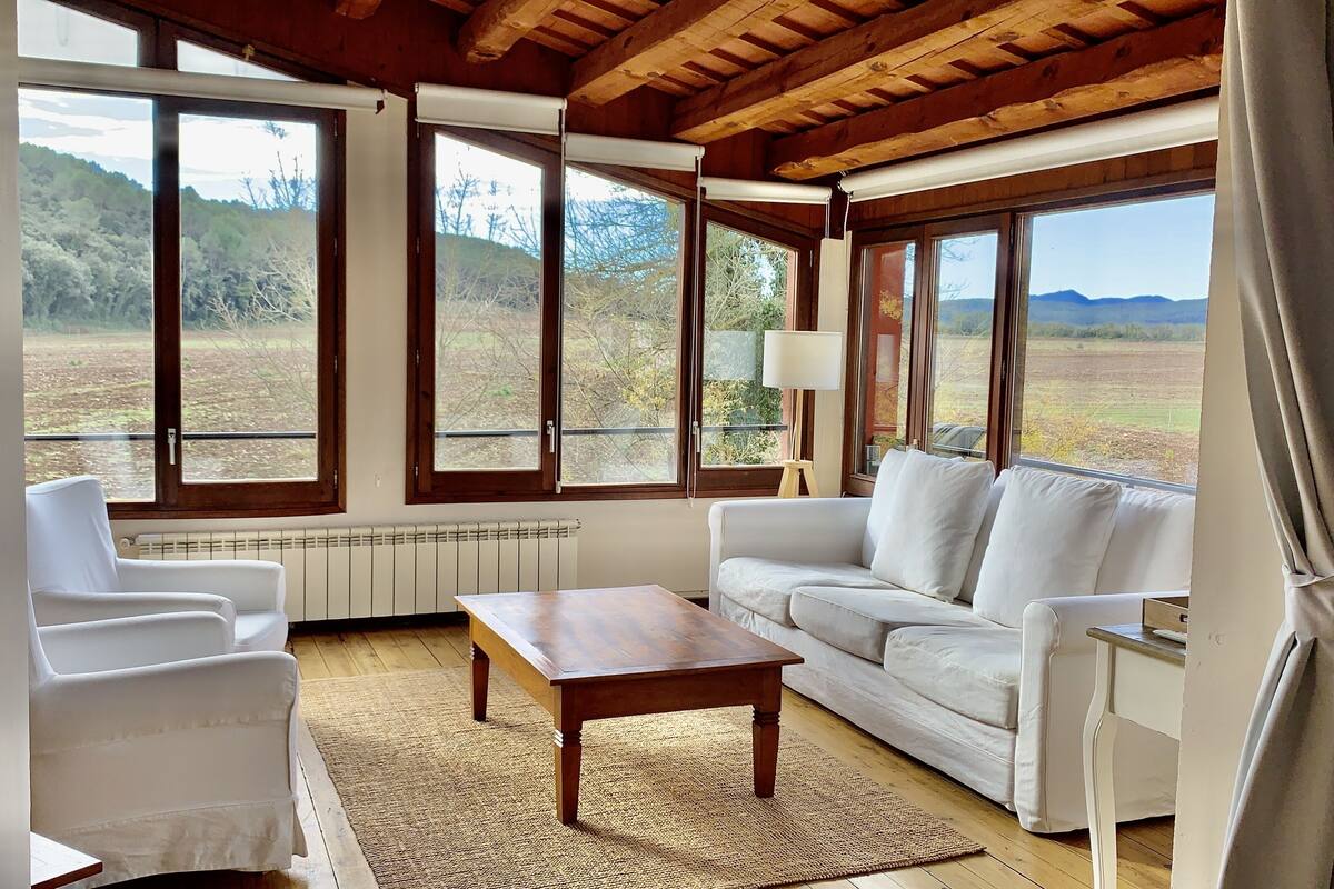 Ein Zimmer mit einem weißen Sofa, einem Holztisch und Blick auf einen Wald durch große Fenster.