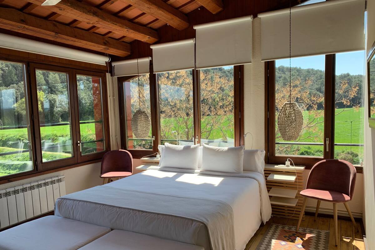 Ein Schlafzimmer mit einem großen Bett, zwei Stühlen und Blick auf eine grüne Landschaft durch große Fenster.