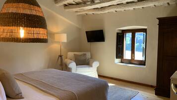 A bedroom with a bed, a hanging light fixture, a floor lamp, a chair, a TV, and a window with shutters.