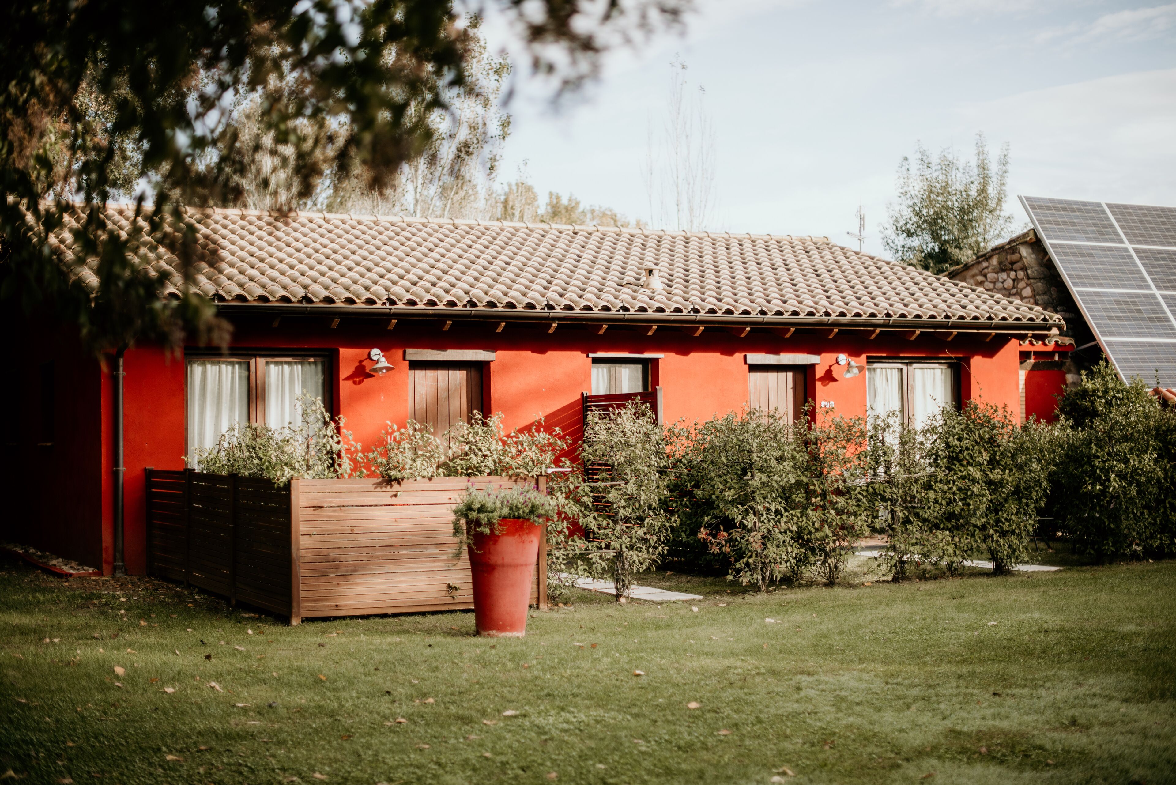 A red building with a tiled roof, wooden shutters, and a garden with a large planter and shrubs.