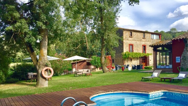A wooden deck with a pool, outdoor furniture, and a stone building in the background. - Hotel Masia la Palma (Cabanelles)