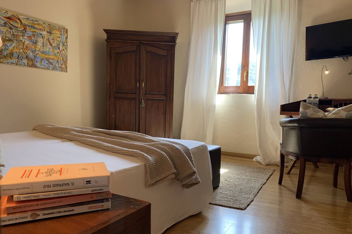 Ein Schlafzimmer mit Bett, einem Holzschrank, einem Schreibtisch mit Stuhl, einem Fenster mit Vorhängen und einem Bild an der Wand.
