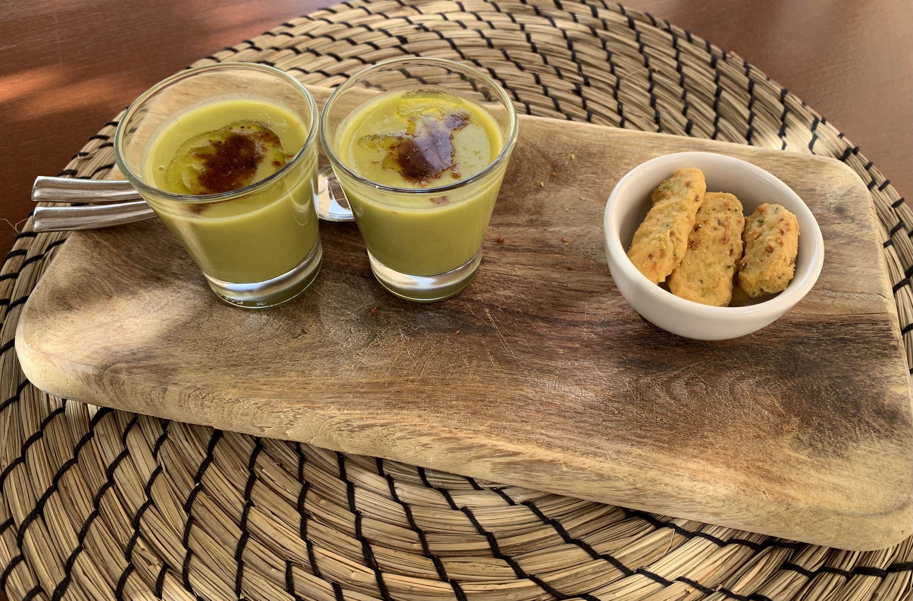 Two glasses of a yellow beverage with a garnish on a wooden board, accompanied by a small bowl of biscuits.