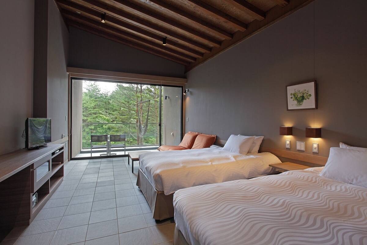 Quarto de hotel moderno com duas camas, televisão e varanda com vista para árvores.