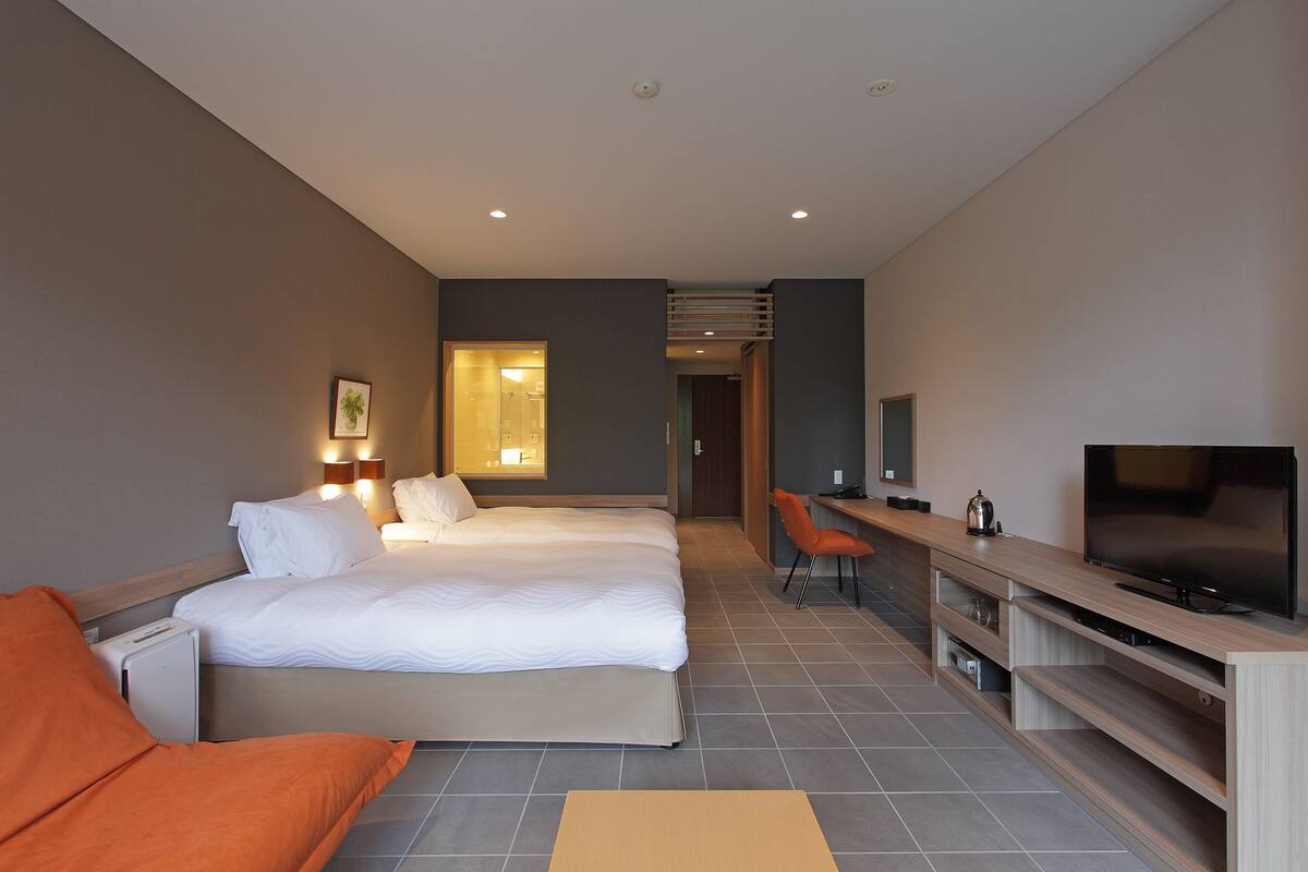 Quarto de hotel com duas camas, uma escrivaninha, uma televisão e um banheiro visível através de uma porta aberta.