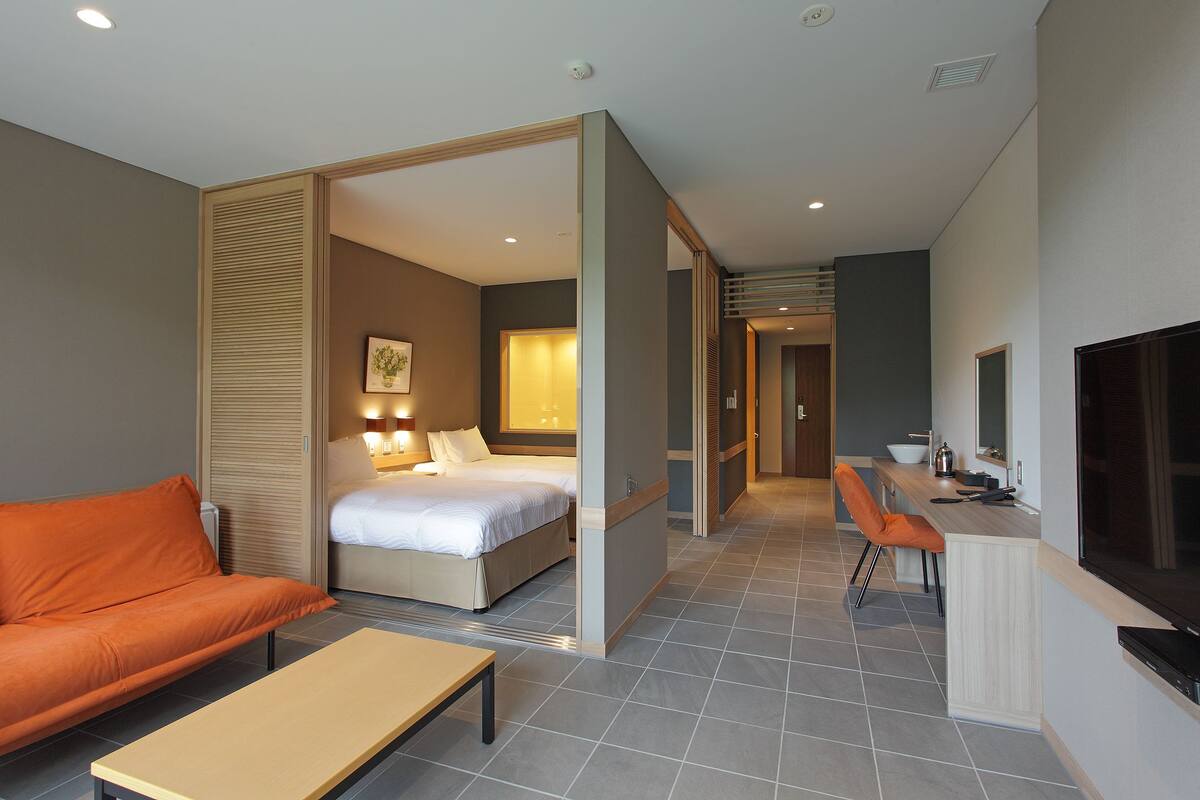 Quarto de hotel com cama, sofá laranja e área de jantar com televisão.