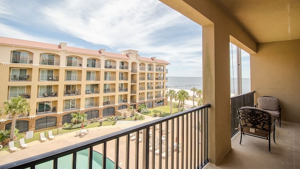 A hotel balcony with a view of the ocean, a pool, and a seating area.