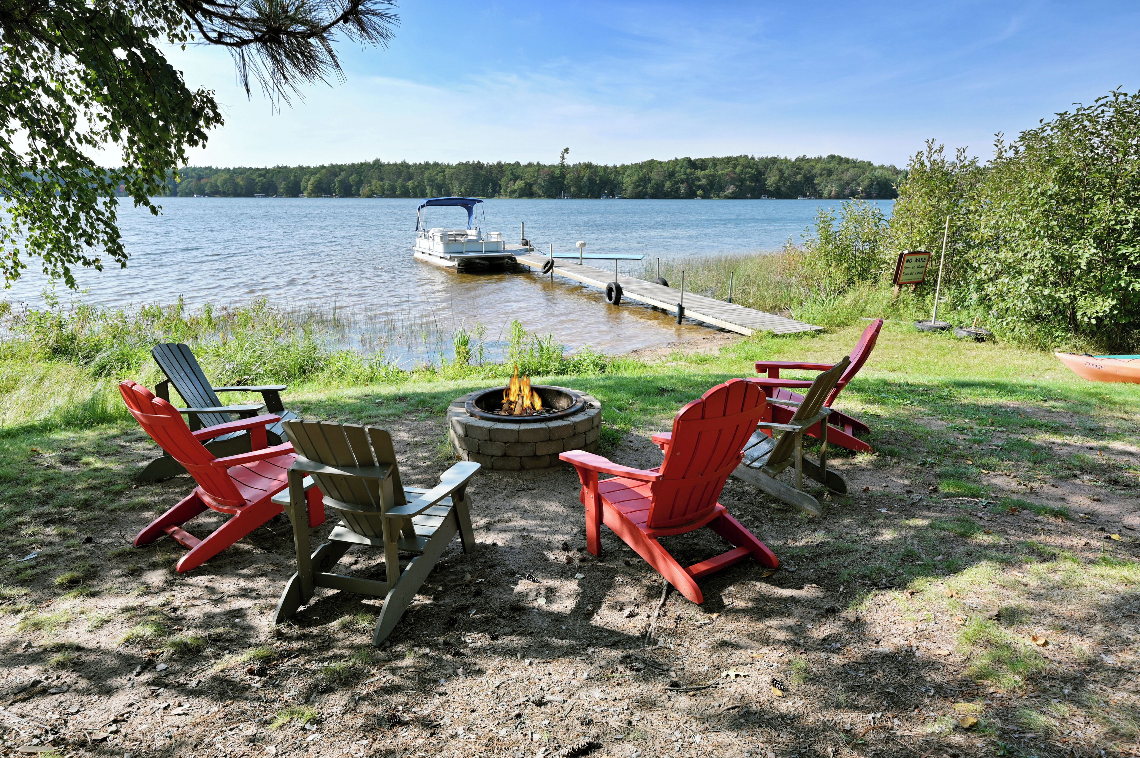 Een terras aan het meer met Adirondack-stoelen, een vuurplaats en een steiger met een boot.