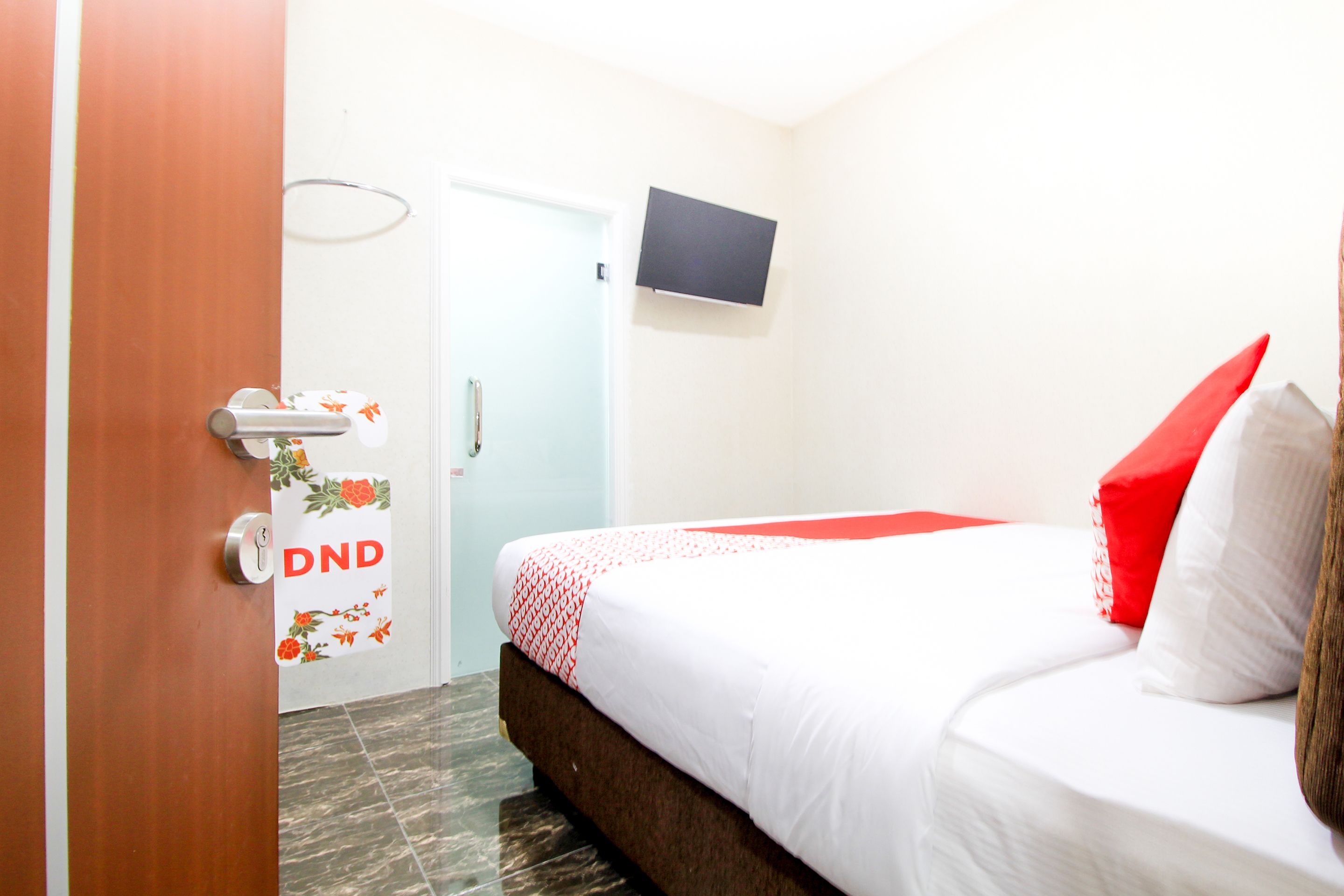 A hotel room with a bed, a television mounted on the wall, a door with a DND sign, and a tiled floor.
