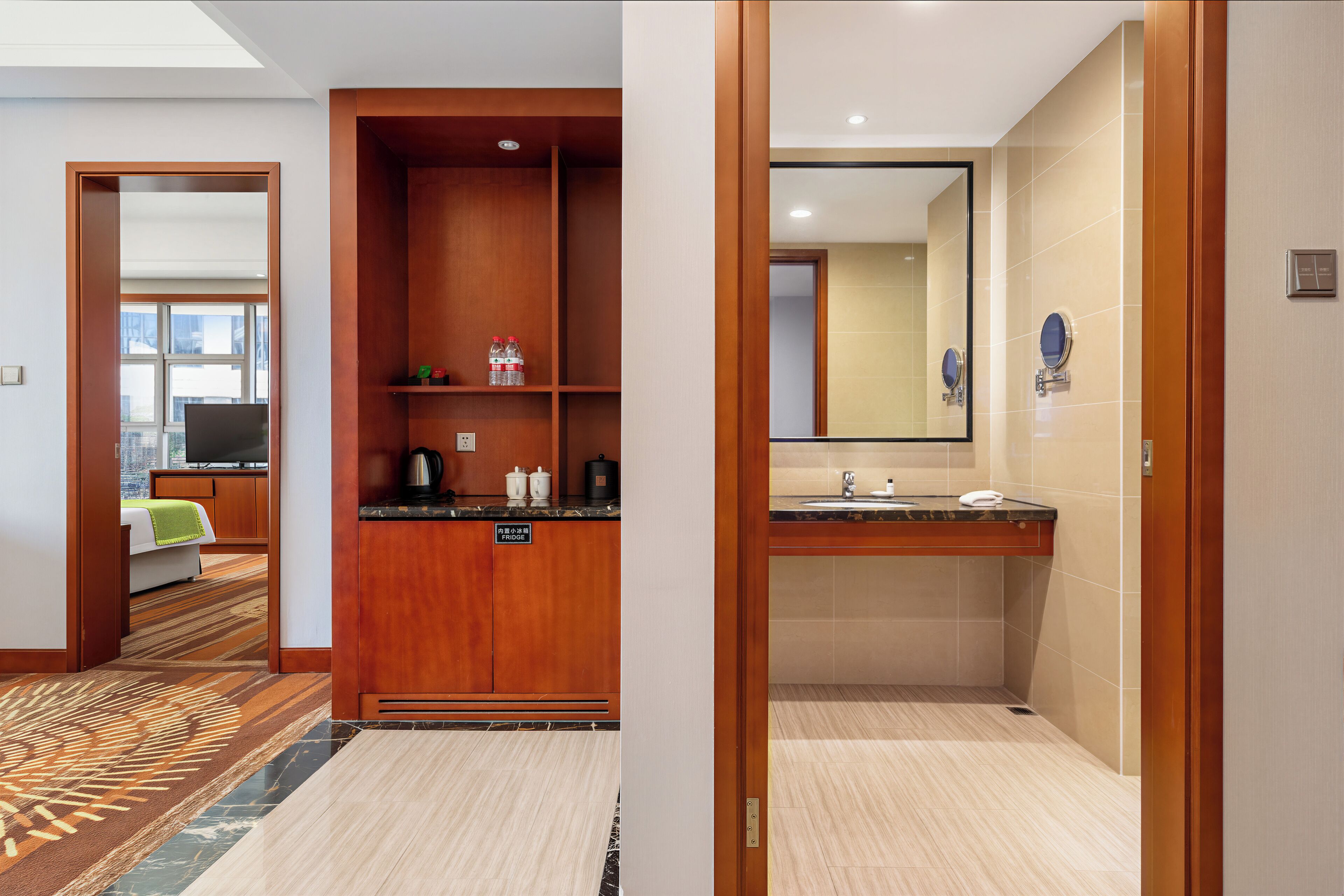 Habitación de hotel con armario de madera, minibar, kitchenette y baño con espejo y lavamanos.