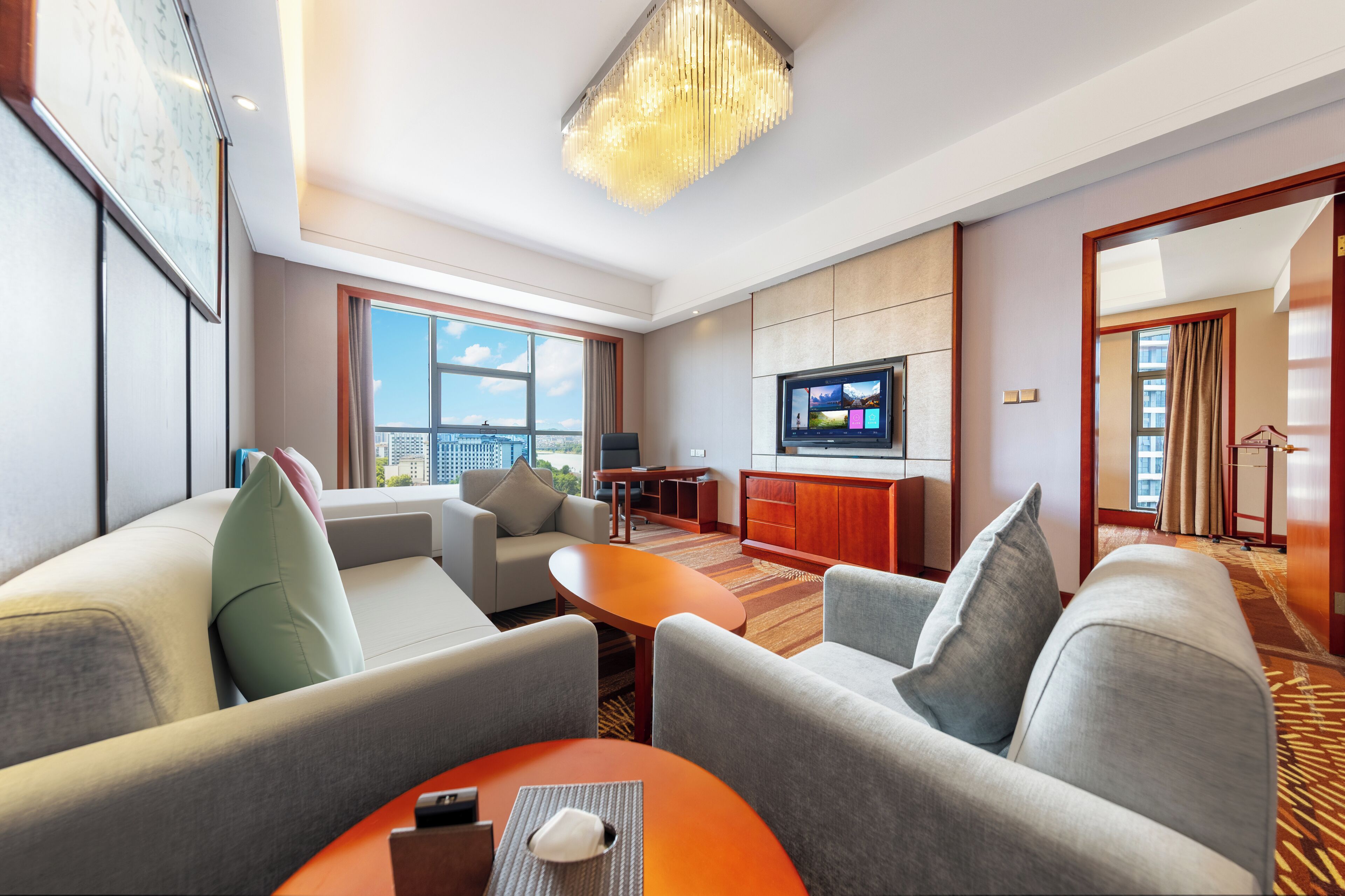 Ein modernes Hotelzimmer mit großem Fenster, Flachbildfernseher, Sofa, Couchtisch und einer Chandelüster.