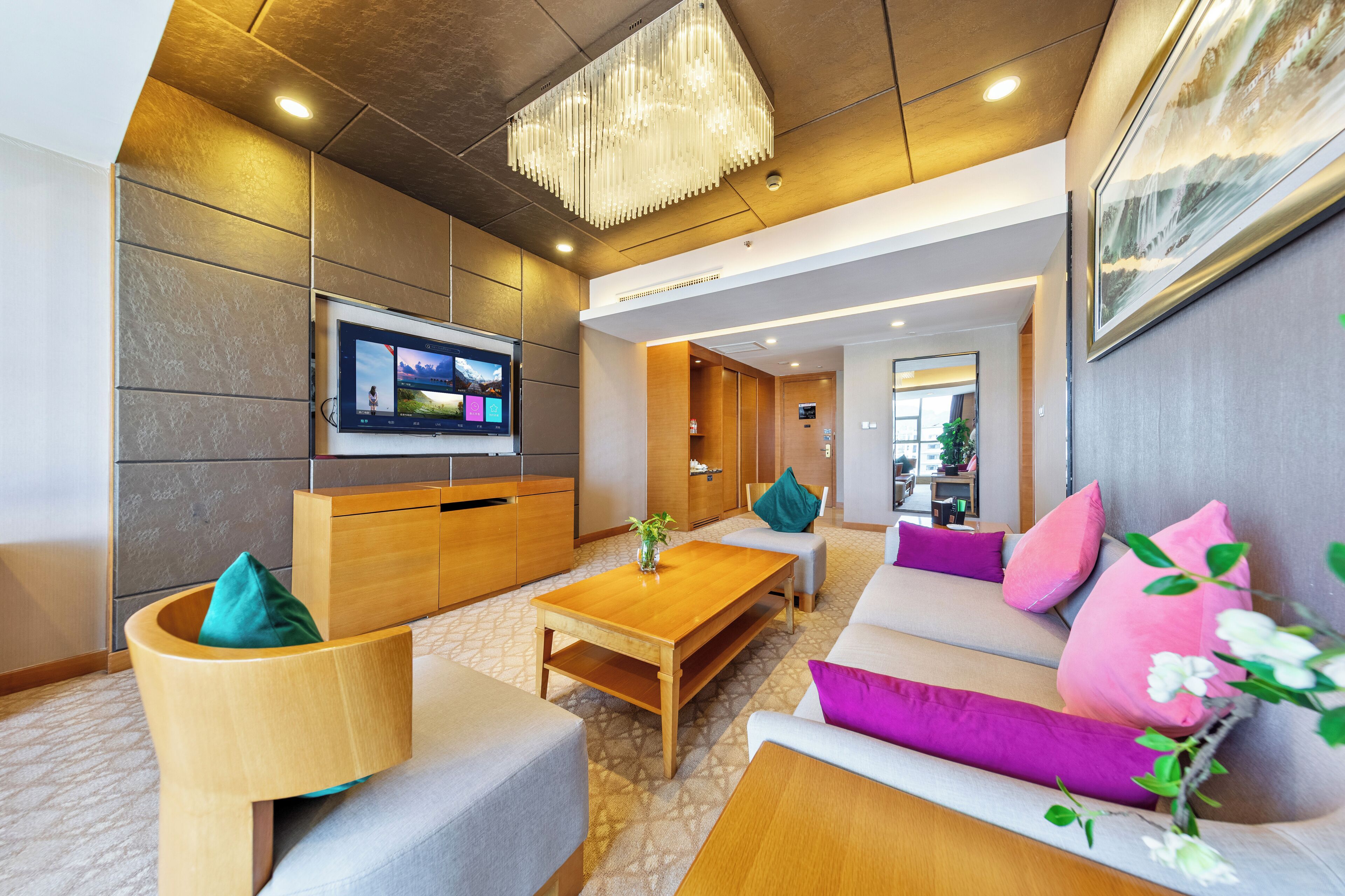 A modern hotel lobby with a large flat-screen TV, wooden furniture, a chandelier, and a painting on the wall.