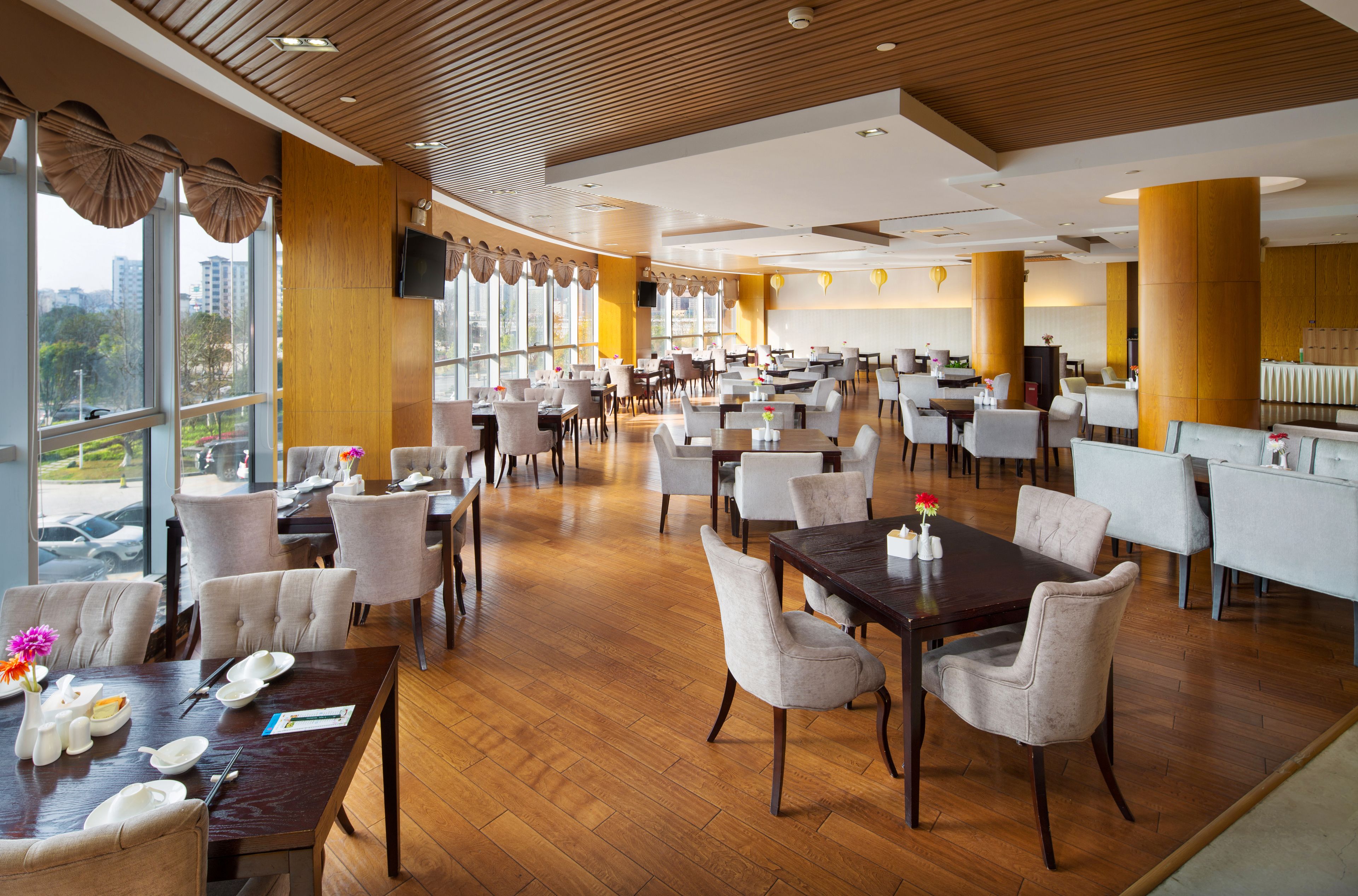 A spacious dining area with multiple tables and chairs, large windows offering a view of the city, and a modern ceiling design.