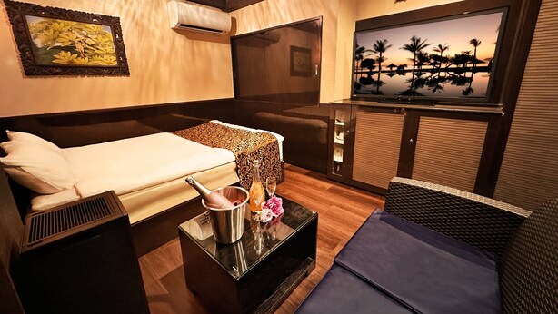 Quarto de hotel com uma cama, uma mesinha com um frasco e flores, uma televisão e vista para palmeiras.