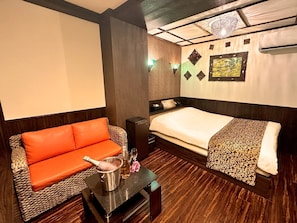 A hotel room with a bed, a sofa, a small table with a bottle and glasses, and a decorative wall piece. - Hotel Gran Bali Resort (Kawasaki)