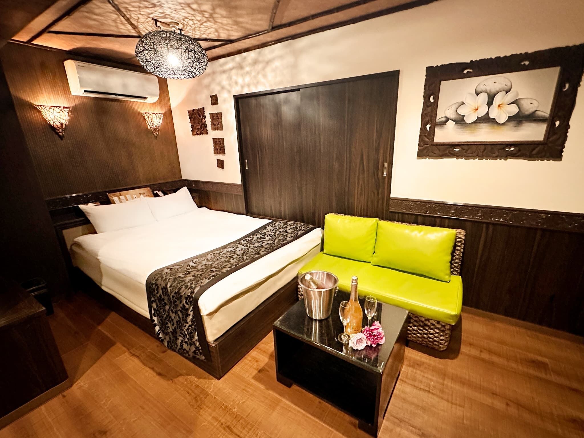 A hotel room with a bed, a green sofa, a small table with a bottle and glasses, and a wall-mounted air conditioner.