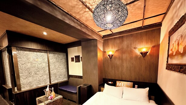 A hotel room with a bed, a nightstand, a lamp, a wall-mounted light, and a decorative wall panel. - Hotel Gran Bali Resort (Kawasaki)