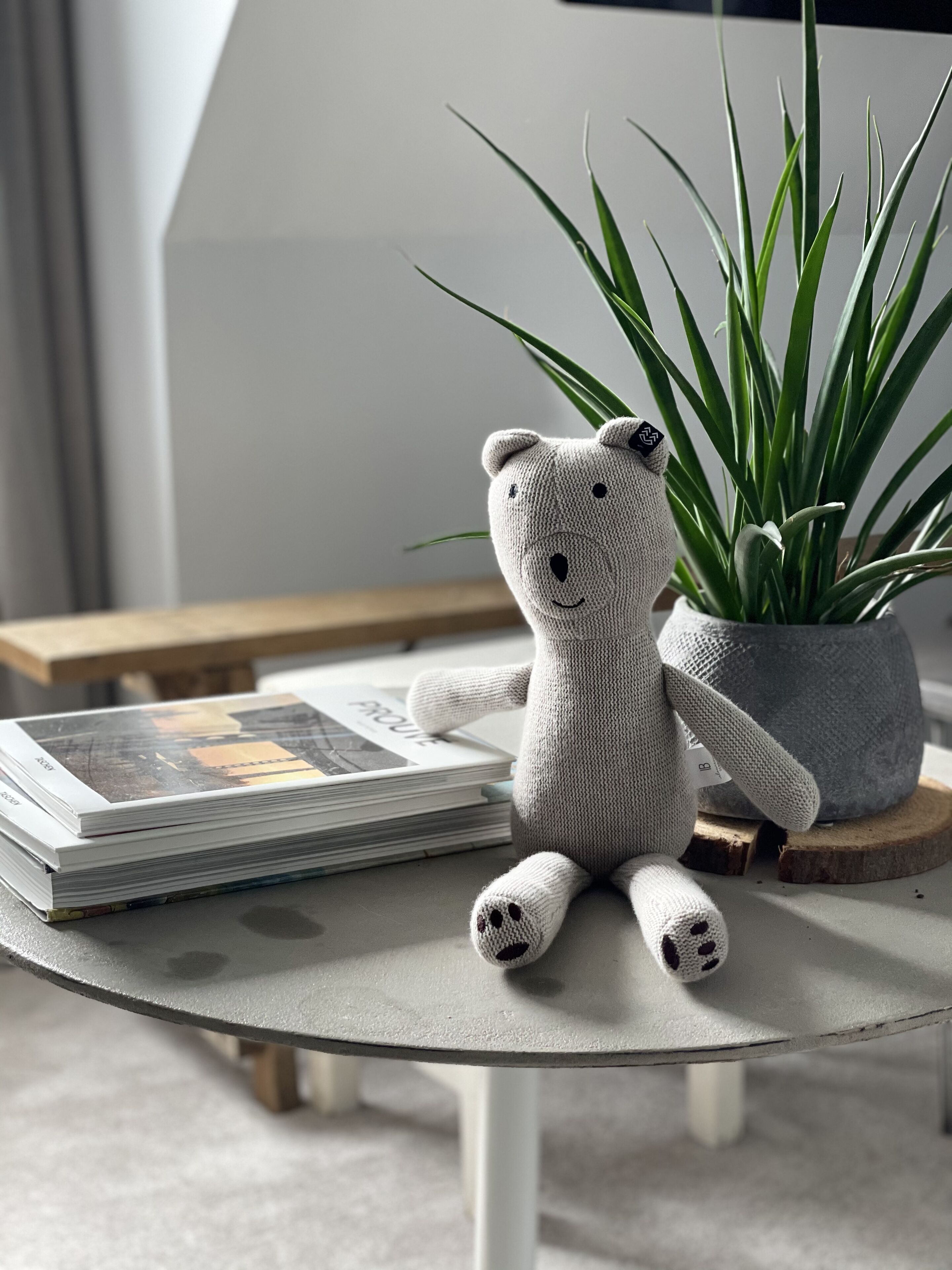 A small knitted bear sitting on a glass table next to a potted plant and a stack of magazines.