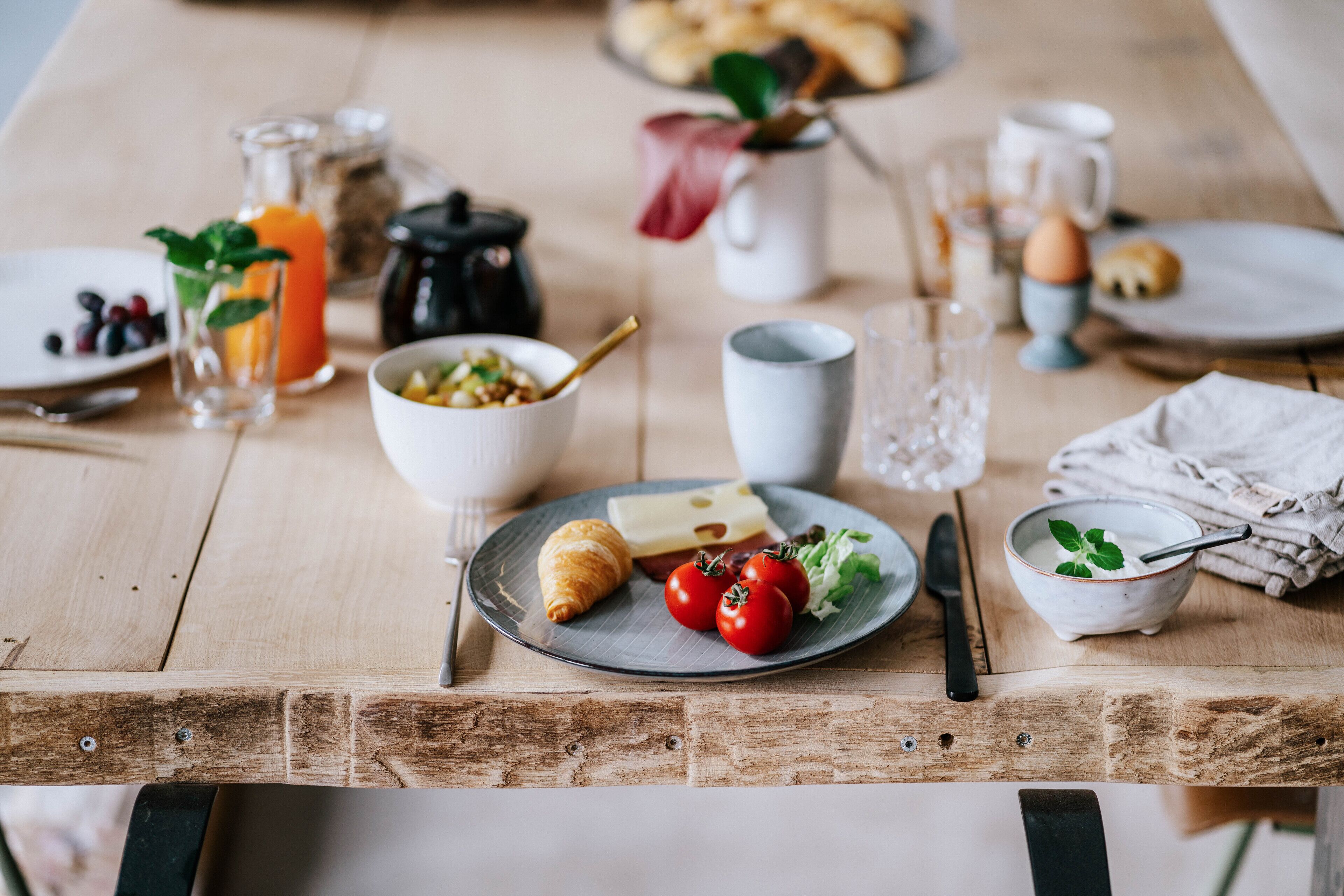 Un desayuno con una mesa de madera, platos, un croissant, tomates, un tazón de cereal, una jarra de jugo de naranja y una cafetera.