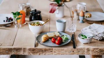 Un desayuno con una mesa de madera, platos, un croissant, tomates, un tazón de cereal, una jarra de jugo de naranja y una cafetera.