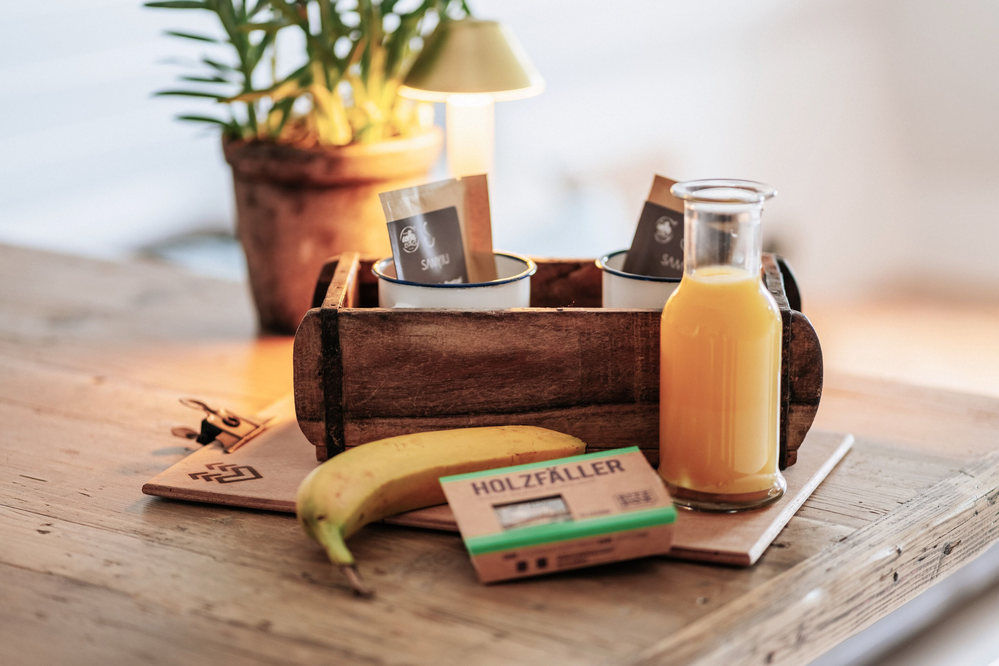 A wooden crate with a glass bottle of orange juice, a banana, and a box labeled "HOLZFÄLLER" on a wooden table.