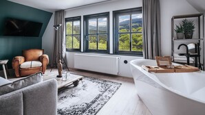 A modern bathroom with a large bathtub, a wooden tray, and a view of a mountainous landscape through large windows. - The Hearts Hotel Boutique und Event Resort (Braunlage)