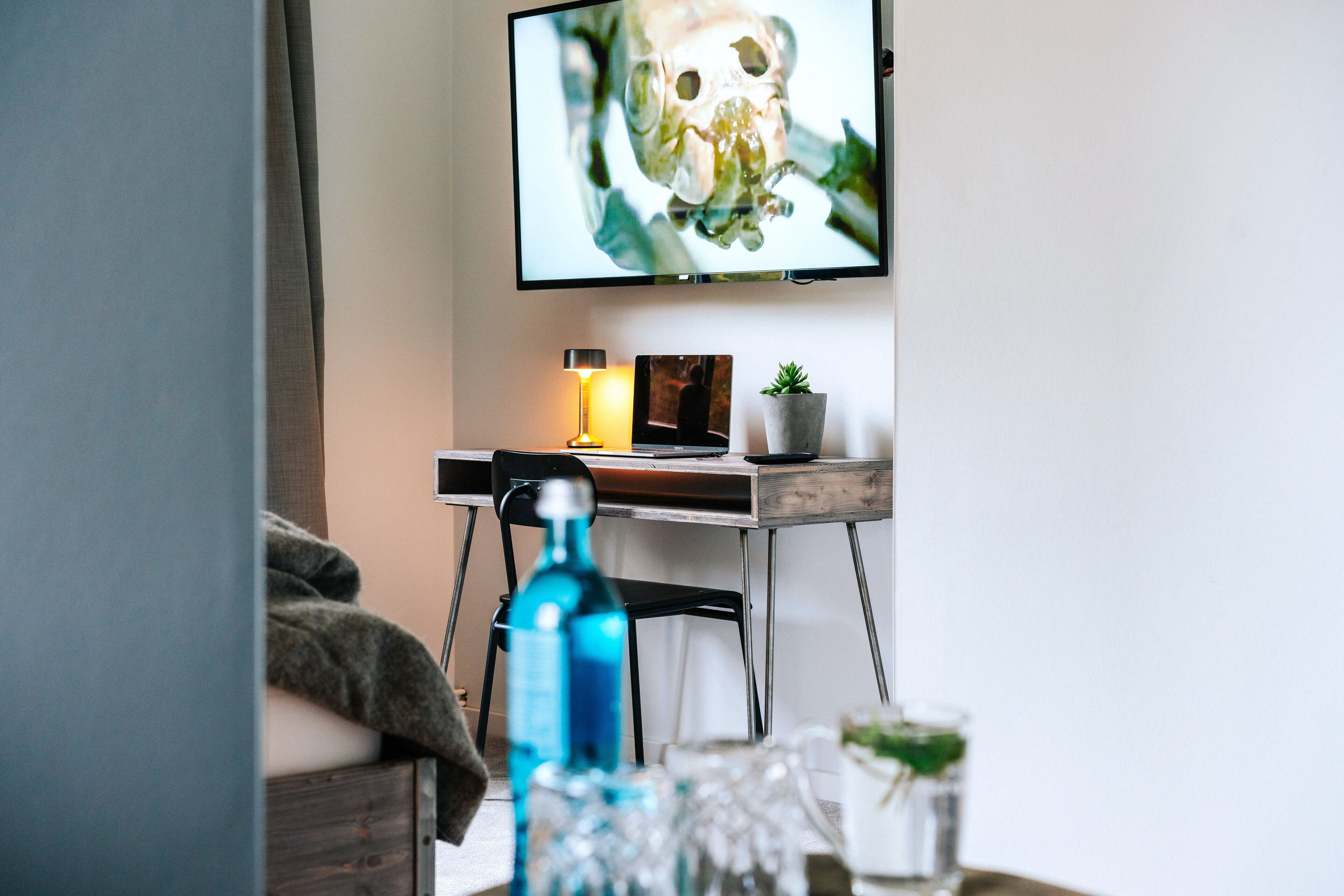 Una habitación moderna con un escritorio, un monitor de ordenador, una lámpara, una planta pequeña y una botella de agua.