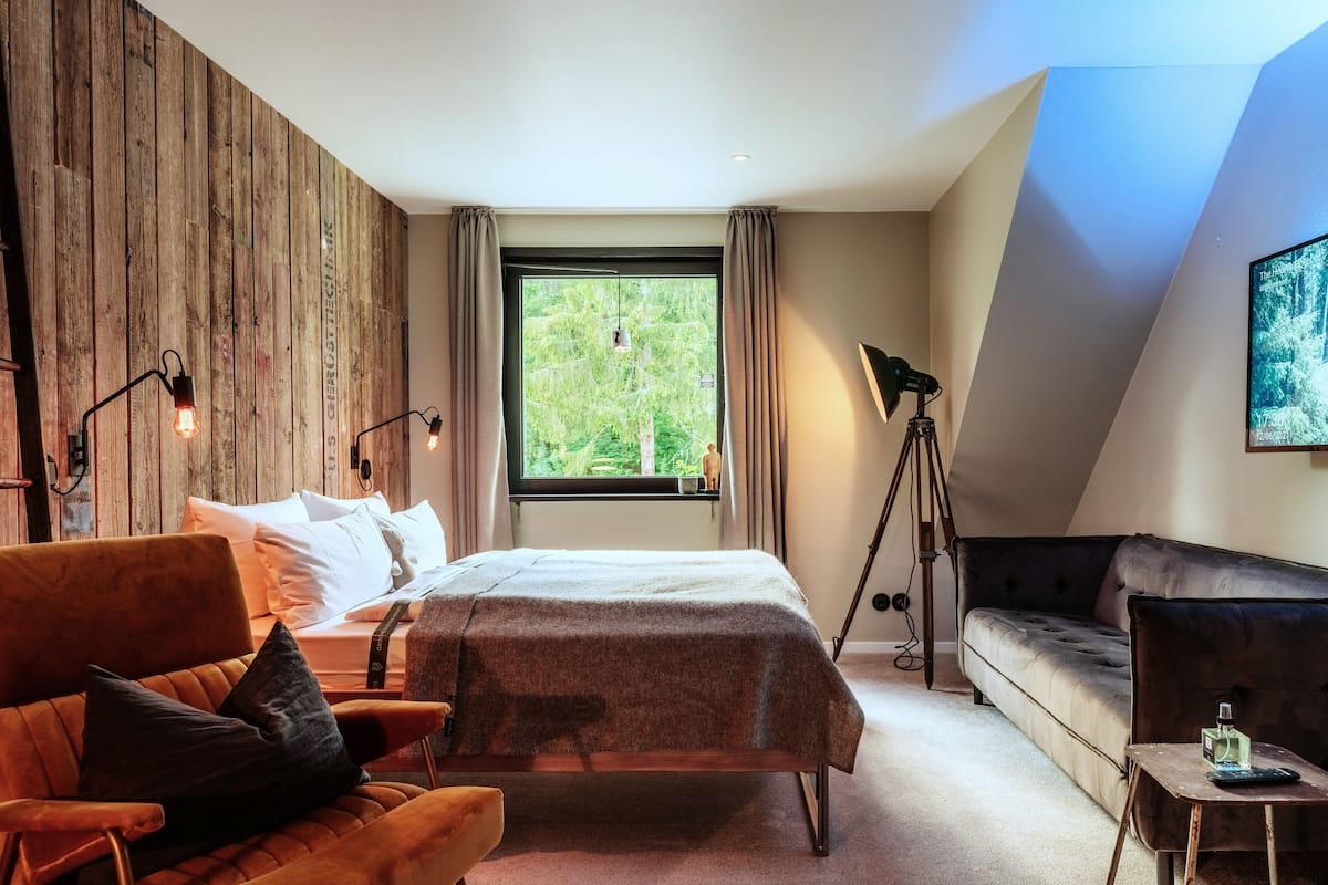 A modern bedroom with a wooden accent wall, a large window with a view of greenery, a bed with a grey bedspread, a brown leather sofa, and a small table with a lamp.