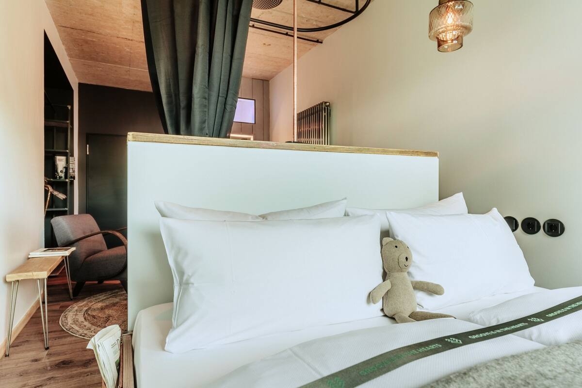 A neatly made bed with white linens and a teddy bear on the pillow, a small side table with a book, a dark armchair, and a modern light fixture.