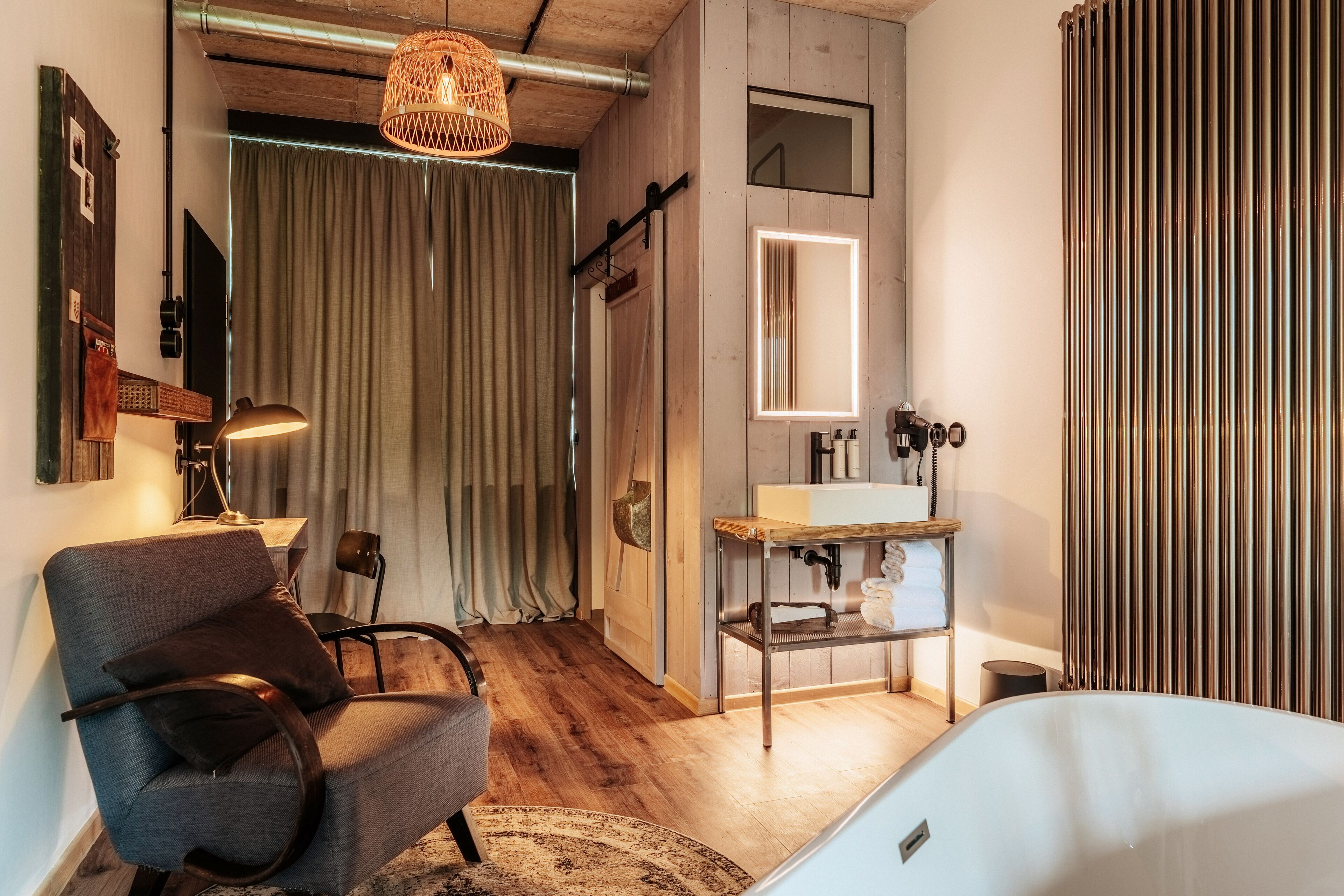 Une chambre moderne avec un plancher en bois, un fauteuil gris, une table de chevet, un lit blanc, un miroir, un lavabo et un rideau.