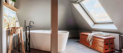 A modern bathroom with a freestanding bathtub, a wooden stool, a painting, and a skylight.